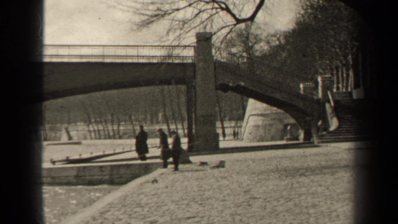 In 1947 Paris, pedestrians in dark coats walk along a riverside embankment near a stone bridge.
