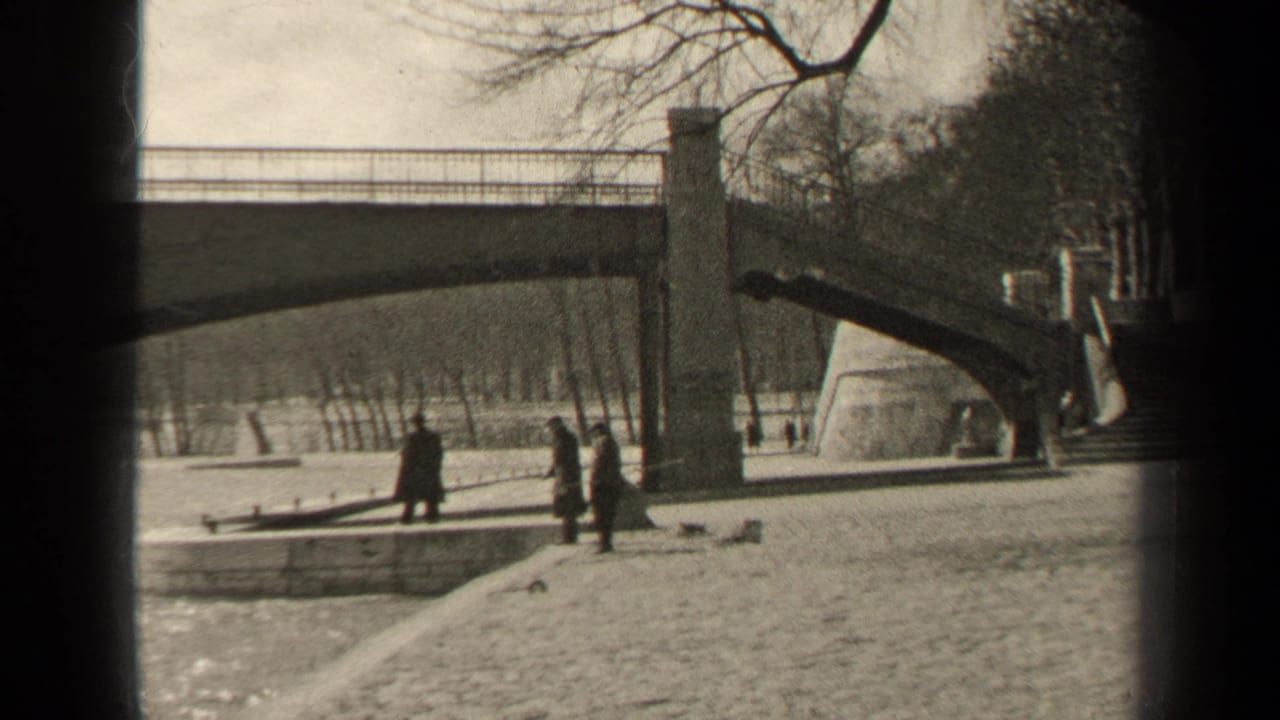 Three figures stand or walk on a riverside path below a bridge.