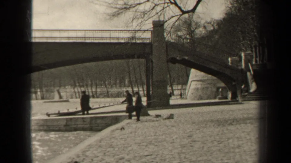 The 1947 Paris home-movie frame shows people in winter coats walking beside water under a stone bridge.