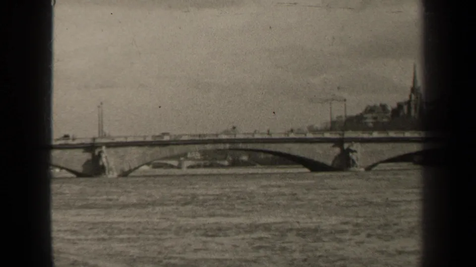 1947 Paris bridge view with cars crossing above the river, a distant church spire, and soft grainy black-and-white 8mm exposure.