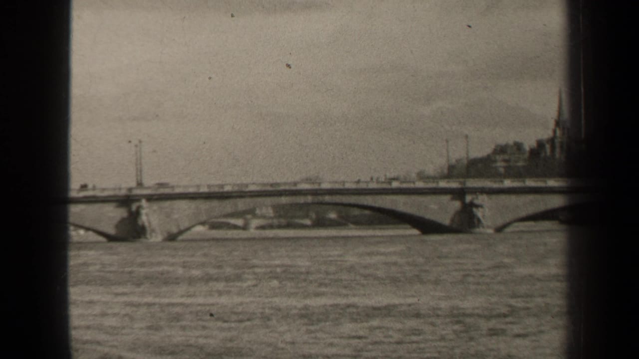 1947 Paris home-movie frame showing a stone arch bridge over a wide river, with vehicles crossing and buildings on the far bank.