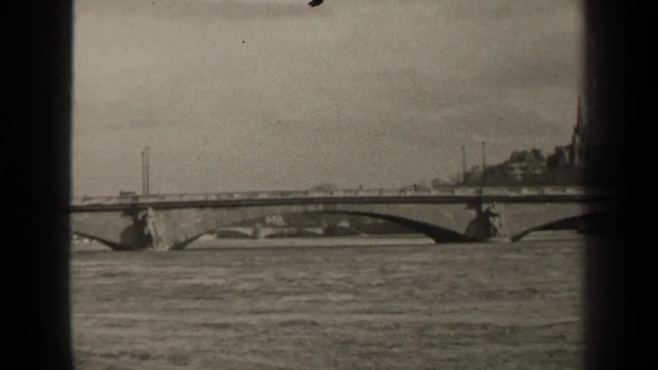 1947 Paris 8mm frame looking across river water toward an arch bridge, with vehicles, distant buildings, and a tall spire visible.