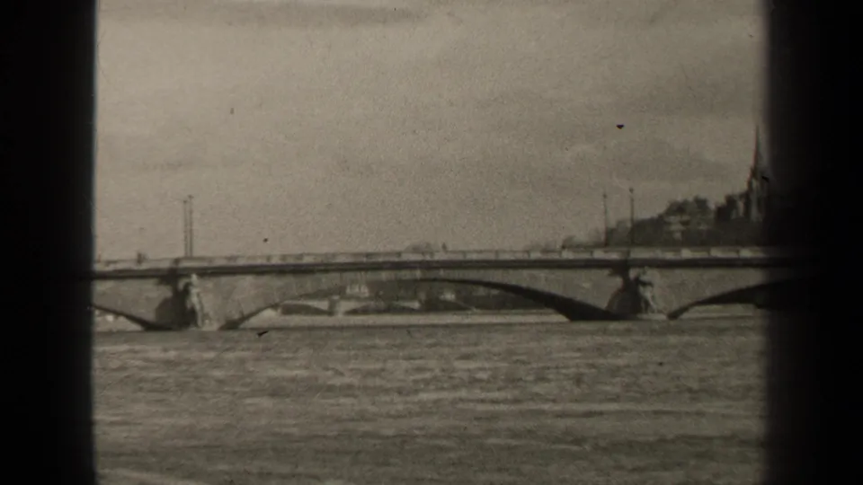 Final 1947 Paris frame showing the river bridge and far-bank buildings, framed by dark 8mm gate edges and soft exposure.
