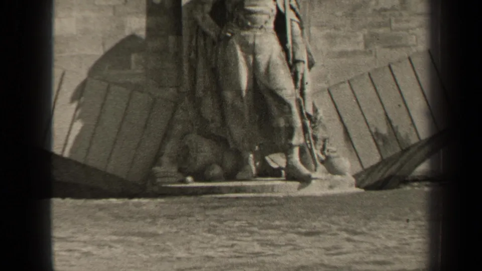 1947 Paris frame focused on the base of a large stone statue by the river, with a wall, planks, and water visible in the foreground.