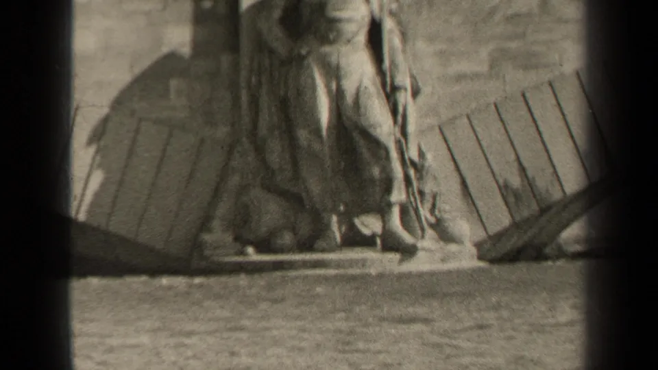 Black-and-white 8mm view of a weathered waterside statue in Paris, with planks, stonework, and waves visible below.