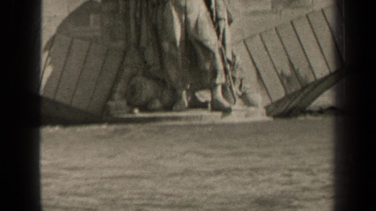 Black-and-white 8mm frame of a Paris monument figure with draped clothing, seen from across water.