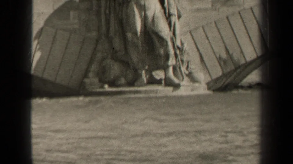 Black-and-white 8mm frame of a Paris monument figure with draped clothing, seen from across water.