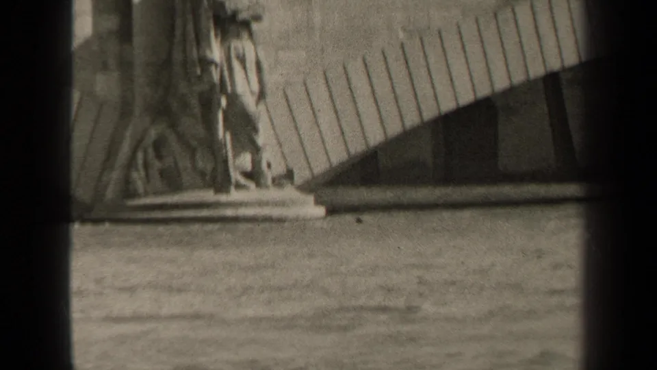 Soft black-and-white home-movie frame of a Paris waterside monument, with a standing figure and curved stone backdrop.