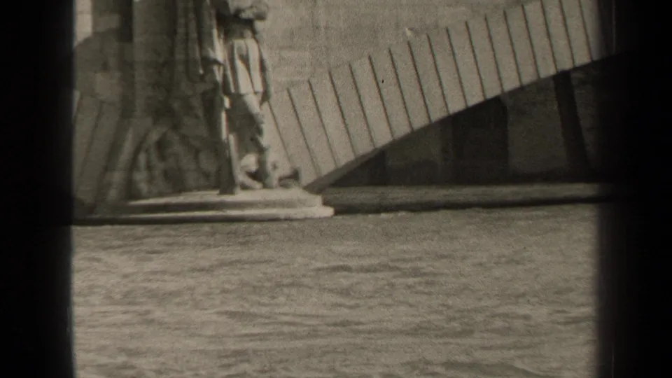 1947 Paris 8mm frame showing a stone statue base near rippling water and a curved slatted structure in the background.