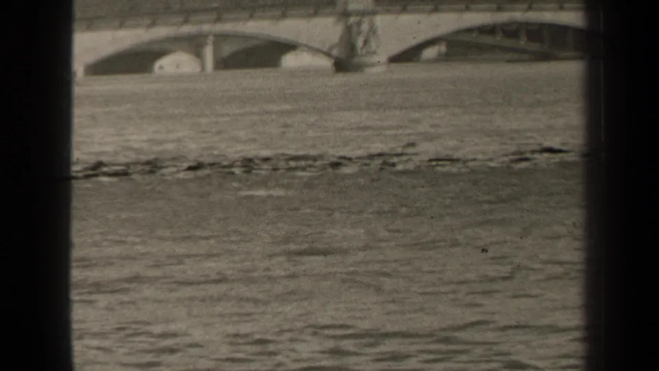 1947 Paris home-movie frame showing churning floodwater below low stone bridge arches, with heavy film grain and gate edges.