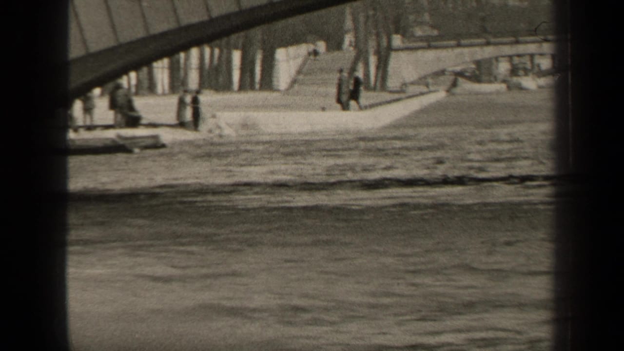Pedestrians on a riverside embankment beneath a bridge with water in the foreground.