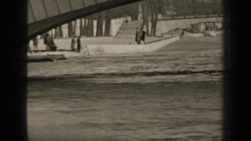1947 Paris riverbank view with small pedestrians walking near the bridge, water in the foreground and trees behind the quay.