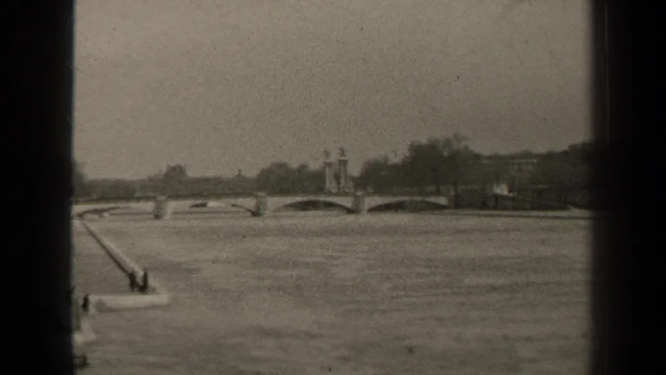 1947 Paris home-movie frame with a broad river foreground, arched bridge beyond, embankment edge, and tiny pedestrians near the water.