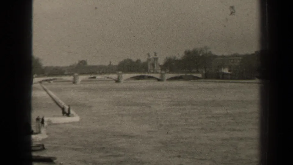 1947 8mm frame of a Paris waterway, showing an arched bridge, riverside embankment, distant pedestrians, trees, and visible film borders.