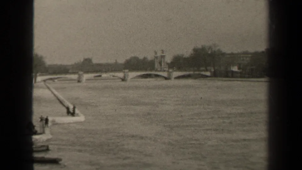 1947 Paris home-movie frame showing a wide river, arched stone bridge, riverside ramp, small boat, and distant figures.