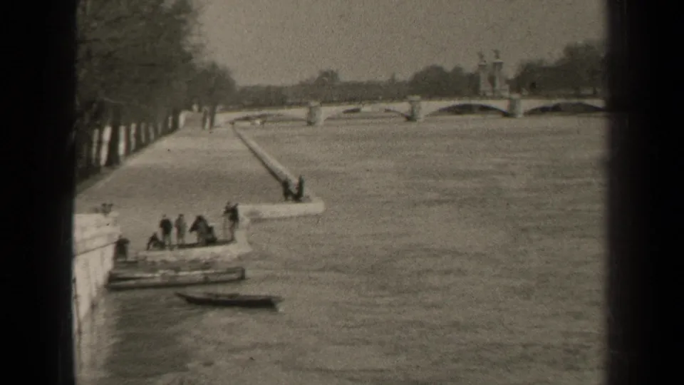 1947 Paris frame with trees along the riverbank, a bridge beyond, a ramp into the river, and people near a small boat.