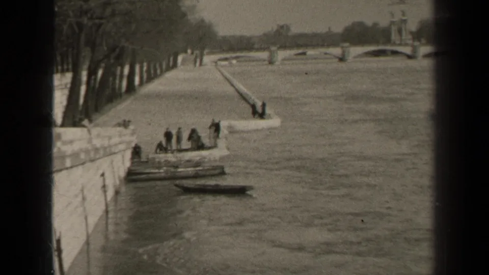 1947 Paris riverside frame showing people beside a small boat at a stone quay, with a ramp, trees, and arched bridge beyond.