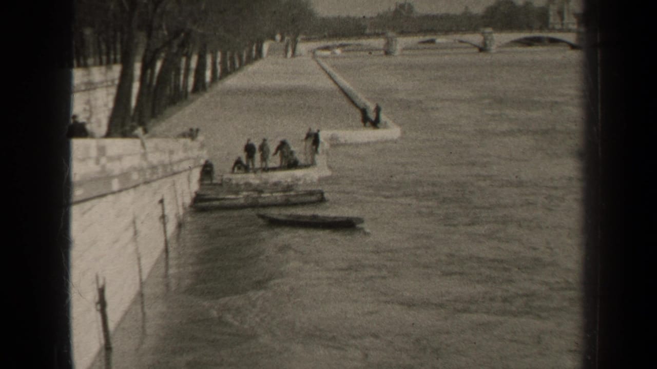 1947 Paris home-movie frame showing people gathered near moored boats beside a stone river quay, with trees and a distant bridge beyond.