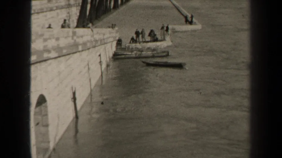 1947 Paris home-movie frame looking along a stone embankment toward people clustered beside moored boats on the river.