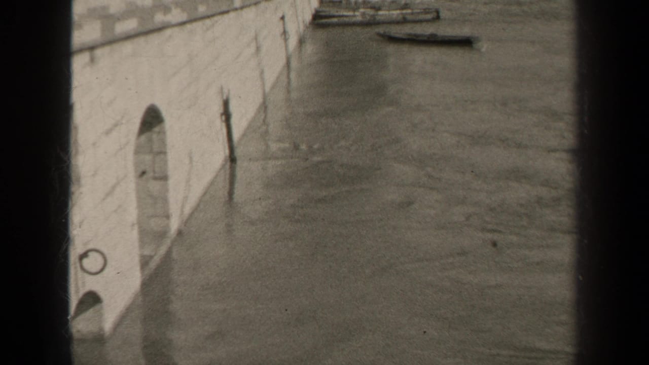1947 Paris floodwater appears to lap against a stone embankment, with small boats visible near the wall in a black-and-white 8mm frame.