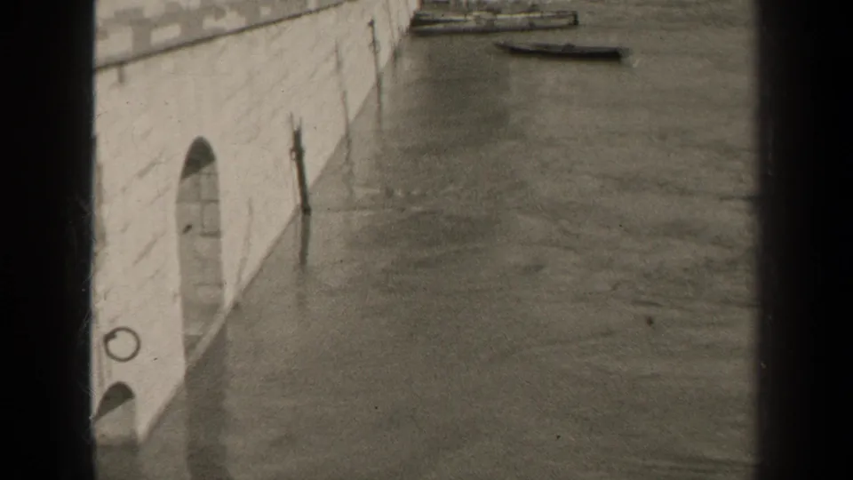 1947 Paris floodwater appears to lap against a stone embankment, with small boats visible near the wall in a black-and-white 8mm frame.