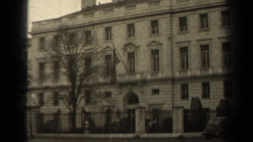 1947 Paris building exterior with a flag above the entrance, bare tree branches, street railings, and small pedestrians near the sidewalk.