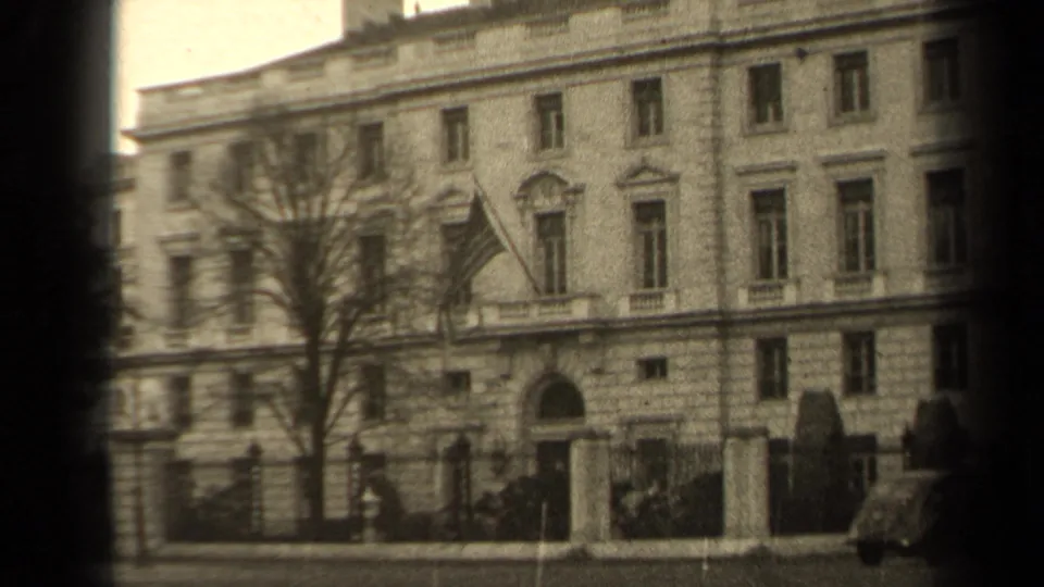 1947 Paris exterior view of a stone building with flag, arched entrance, sidewalk railings, bare tree, and an incidental pedestrian at the edge.