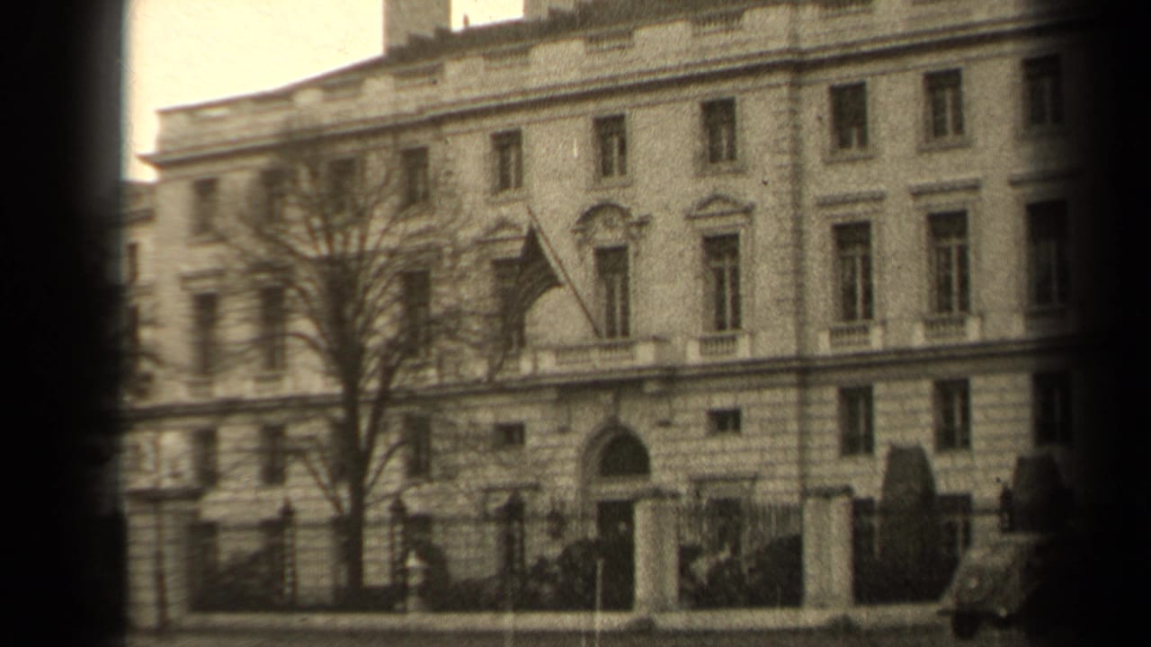 1947 Paris home-movie frame showing a classical stone building, bare tree, iron fence, gate posts, and a flag over the entrance.