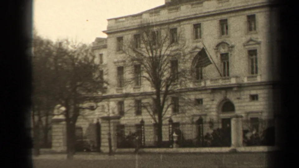 1947 Paris home-movie still showing the building entrance, flag, leafless tree, iron fence, gate posts, and parked cars in soft 8mm focus.