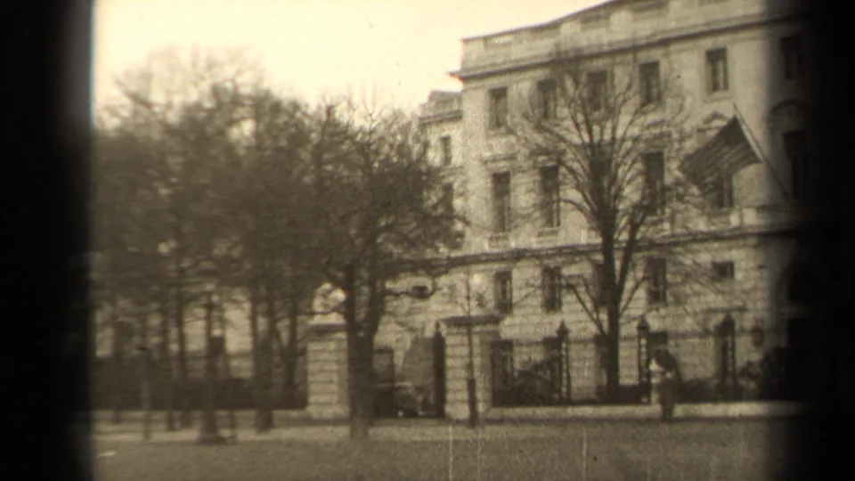 Soft-focused 1947 Paris street footage showing the building facade, bare trees, low fence, pedestrians, and U.S. flag.