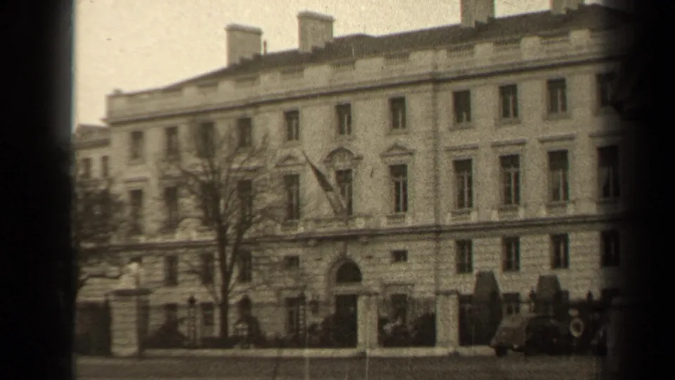 1947 Paris frame showing the building entrance, U.S. flag, fence line, bare tree, and parked cars near the curb.