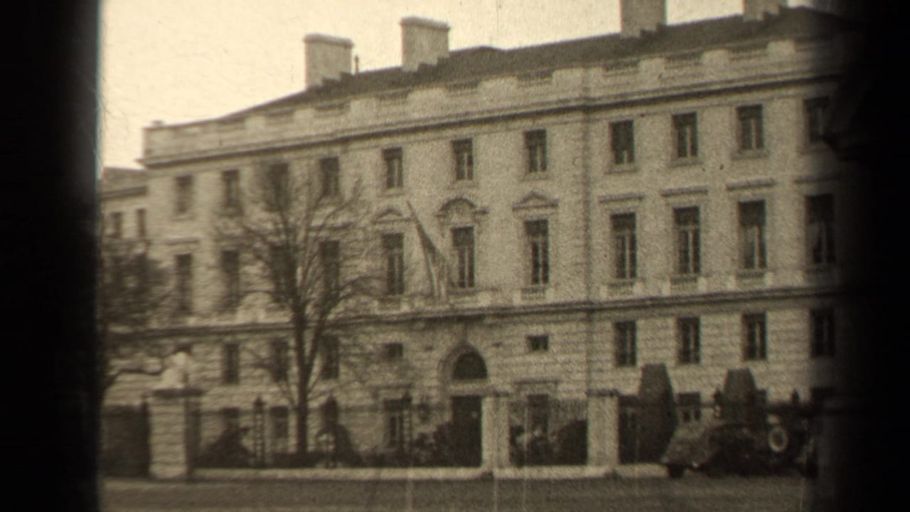 Large pale building facade with central arched doorway, bare tree, iron fence, and cars near the curb.