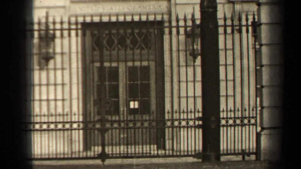 1947 Paris home-movie frame focusing on an ornate iron gate, double doors, stone columns, and the entrance beyond.