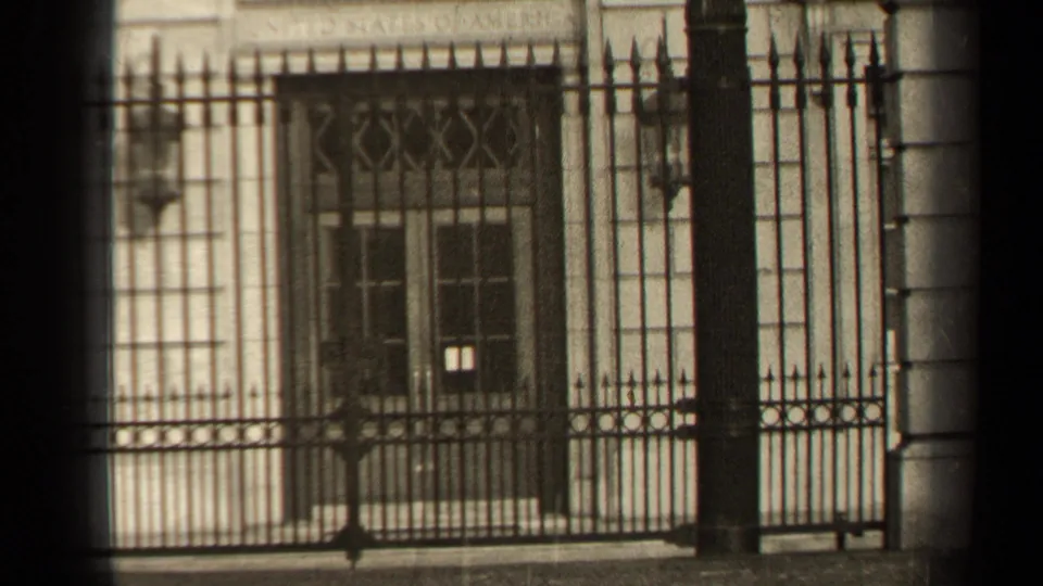 1947 Paris home-movie frame showing a pale stone building behind ornate iron railings, with barred windows and soft winter light.