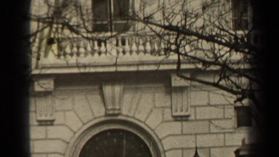 1947 Paris 8mm frame showing a pale stone facade, arched window, balcony balustrade, bare branches, and dark vertical foreground bands.
