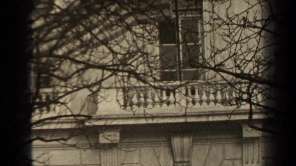 1947 Paris 8mm frame of a stone balcony facade seen through bare winter branches, with a dark vertical foreground band near center.