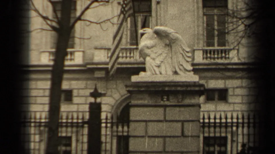 1947 Paris frame with the stone eagle centered above a gate pillar, backed by iron railings, windows, and bare trees.