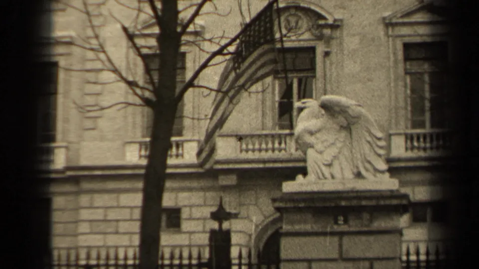1947 Paris 8mm frame showing a U.S. flag extending from a window above a stone eagle statue, with bare branches and an iron fence below.