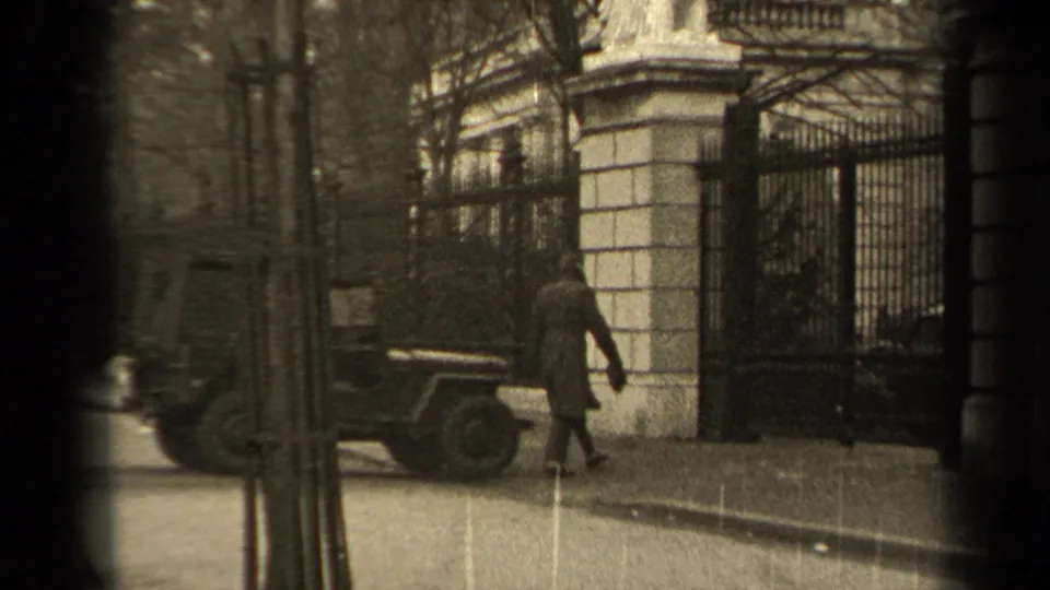 1947 Paris 8mm frame showing a man in a long coat walking on the sidewalk toward a wrought-iron gate near a parked jeep.