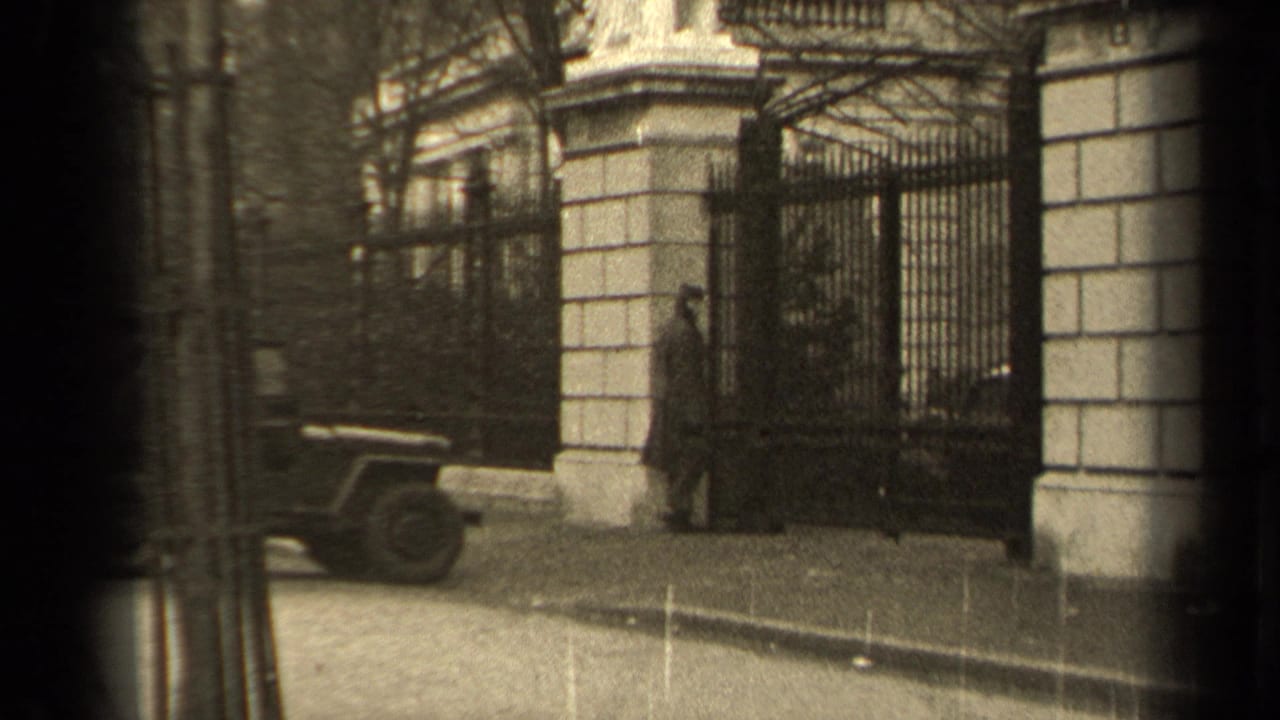 1947 Paris street view with a man in a long overcoat near a large iron gate, stone pillars, and a parked jeep.