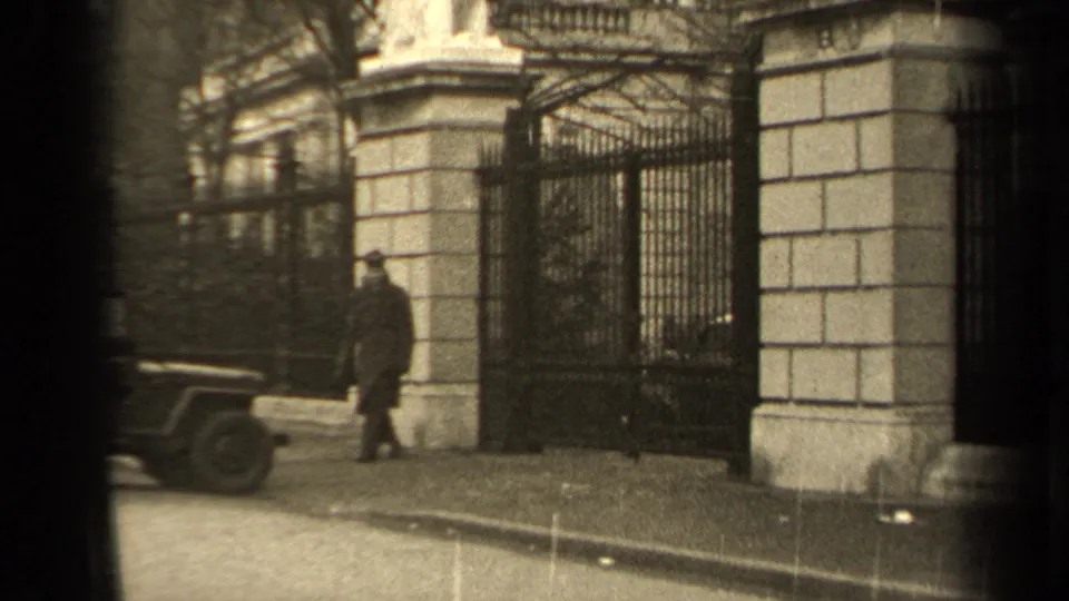 The man in a long coat moves along a Paris sidewalk toward a grand iron gate and stone pillar, with a jeep at the curb.