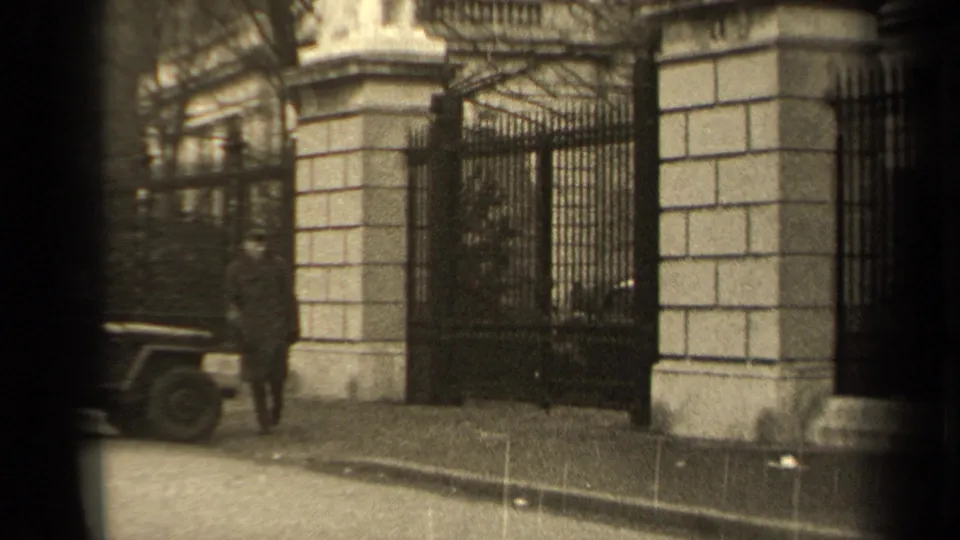 A man in a dark coat stands near a parked jeep outside a large iron gate and stone building in 1947 Paris footage.