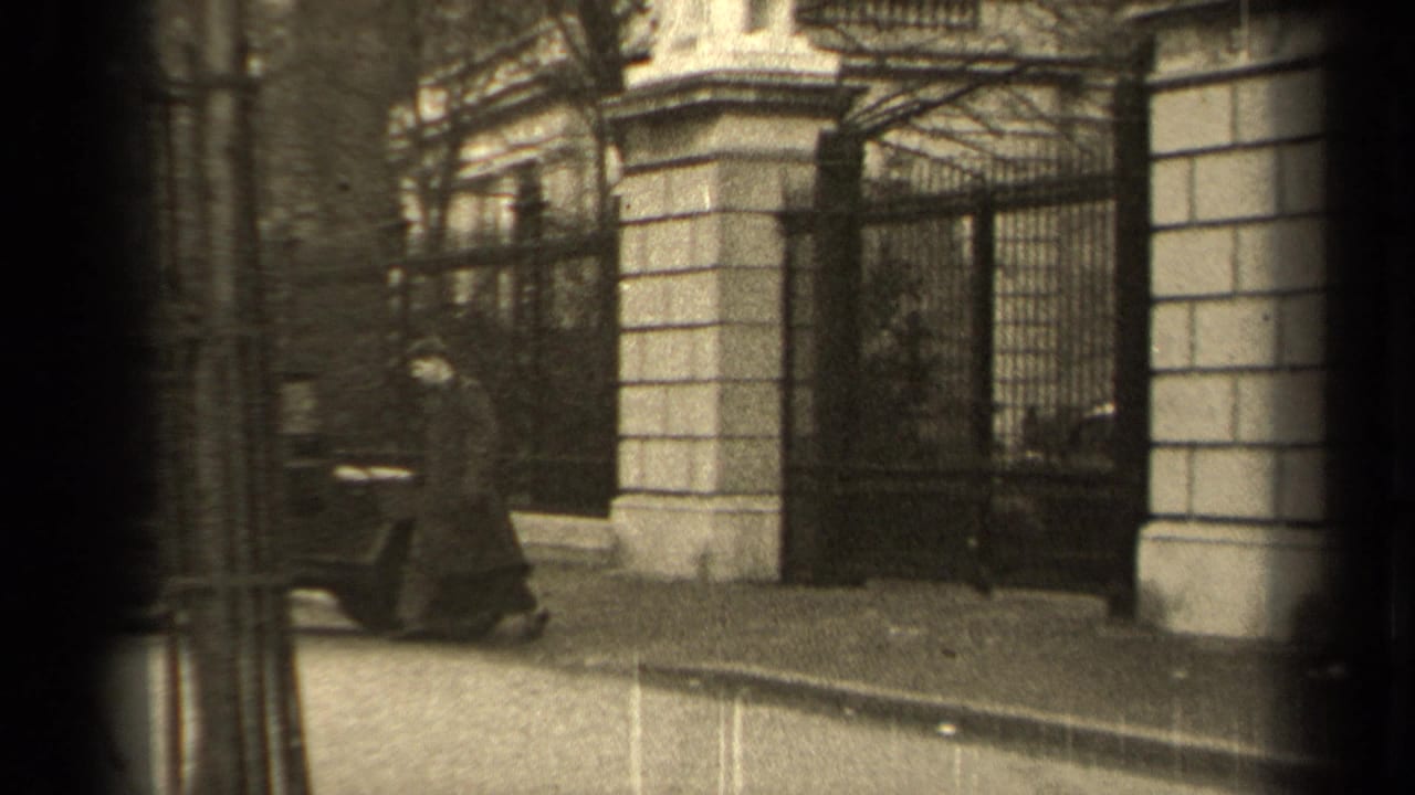 In 1947 Paris footage, a man in a long coat walks toward a guarded iron gate beside a stone facade.