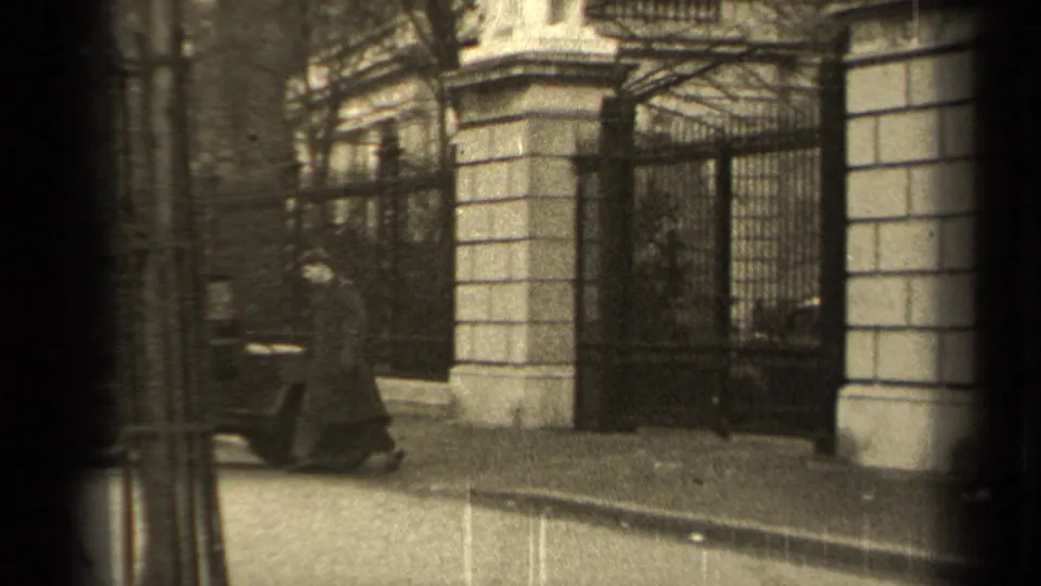 In 1947 Paris footage, a man in a long coat walks toward a guarded iron gate beside a stone facade.