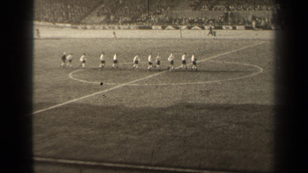 Football players in light tops and dark shorts move in formation across a Paris pitch, with a crowd visible beyond the field.