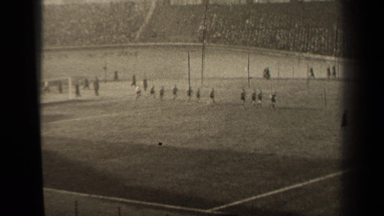 Players walk across the soccer pitch while spectators fill the background stands.