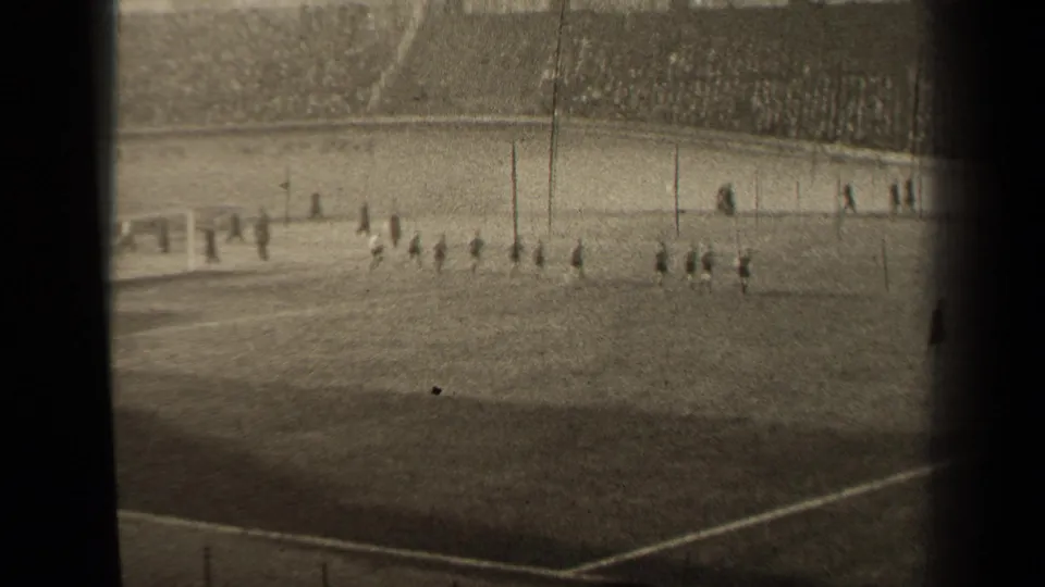 Distant black-and-white 8mm view of soccer players crossing the pitch in the stated 1947 Paris stadium setting.