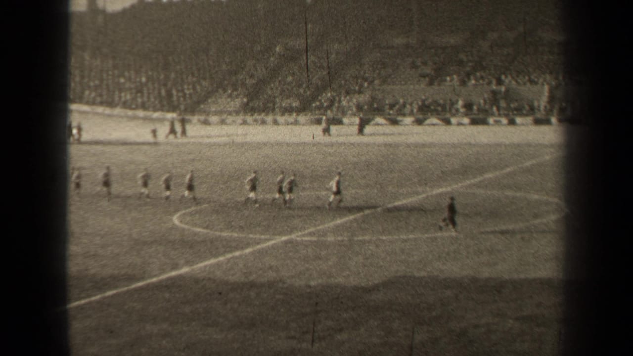Soccer players jog across the field while spectators fill the background stands.