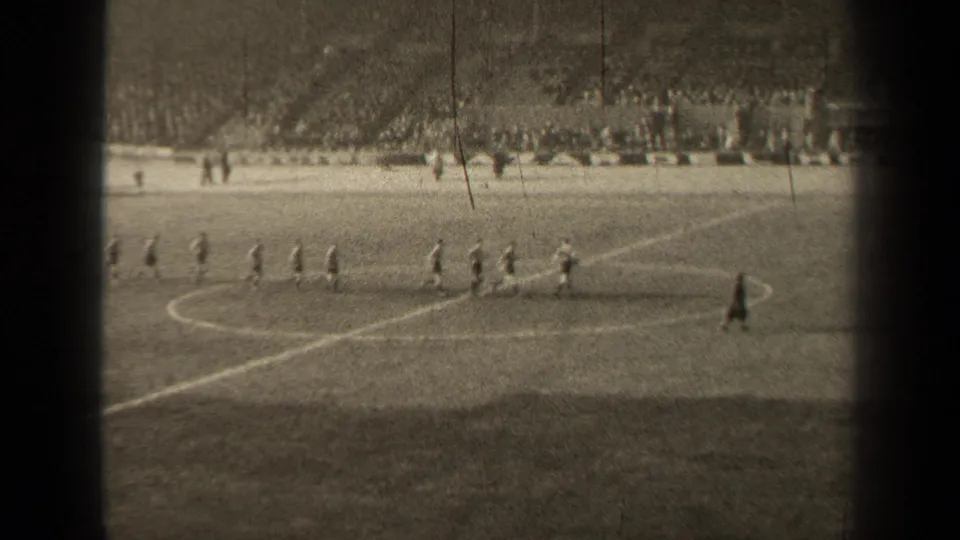 Distant 1947 Paris stadium view of soccer players crossing the center-circle area with spectators and sideline figures behind them.
