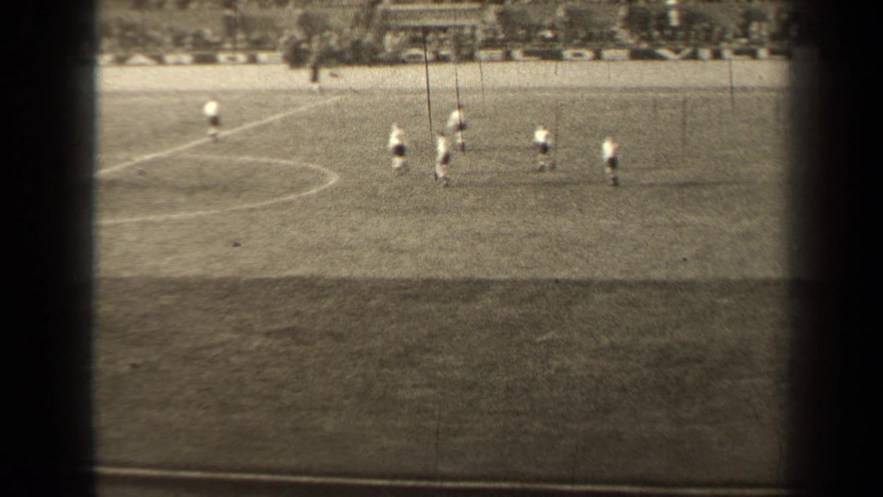 1947 Paris 8mm frame showing distant soccer players spread across a marked grass pitch, with spectators faintly visible beyond the field.